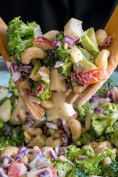 broccoli salad being held up by a wooden spoon with dressing on it and other vegetables in the background