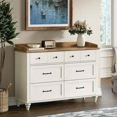 a white dresser with flowers in a vase and a painting on the wall above it