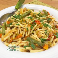 a white bowl filled with pasta and vegetables on top of a wooden table next to a spoon