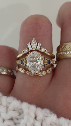 a woman's hand with two wedding rings on top of her finger and the ring has an oval shaped diamond surrounded by smaller round diamonds