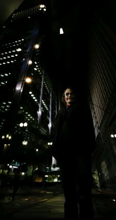 a man standing in front of a tall building at night with the lights on and buildings lit up behind him