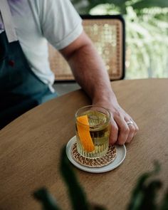 a man holding an orange slice on top of a glass cup filled with tea sitting on a wooden table