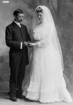 an old black and white photo of a man and woman dressed in wedding attire standing next to each other