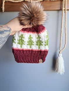 a hand holding a knitted hat on top of a wooden shelf with tassels hanging from it