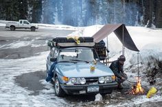 a car is parked in the snow next to a campfire with two people cooking on it