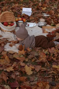 a small child laying on the ground with leaves around him and apples in a basket