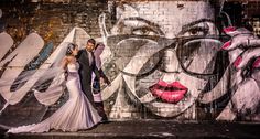 a bride and groom standing in front of a graffiti wall with their veil blowing in the wind