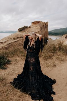 a woman standing on top of a sandy beach next to the ocean wearing a black dress