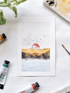 watercolors and paint supplies sit on a table next to an art work with a house in the background