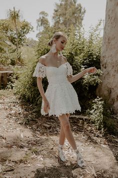 a woman in a white dress standing next to a tree and bushes with her hand on her hip