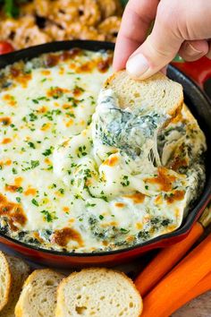 a hand dipping a piece of bread into a skillet filled with spinach and cheese