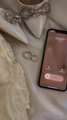 a cell phone sitting on top of a white bed next to wedding shoes and rings