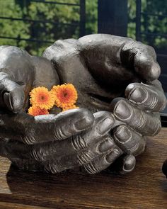 a hand holding an orange flower sitting on top of a wooden table next to a window