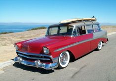 an old red station wagon with a surfboard on the roof is parked by the ocean