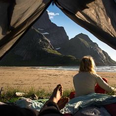 a woman sitting on top of a bed under a tent next to the ocean with mountains in the background