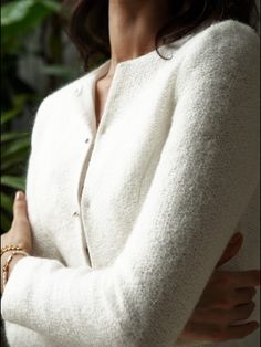 a woman with her arms crossed wearing a white sweater and gold bracelets, standing in front of green plants