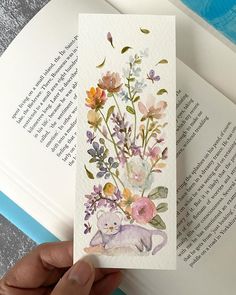 a hand holding an open book with watercolor flowers on it and a cat in the middle