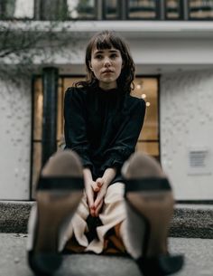 a woman sitting on the ground in front of a building with her legs crossed and looking at the camera