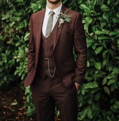 a man wearing a suit and tie standing in front of some bushes with his hands in his pockets