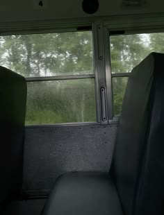 an empty seat in the back of a bus on a rainy day with trees and grass behind it