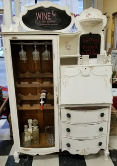 an old fashioned white cabinet with wine glasses on the top and bottles in the bottom