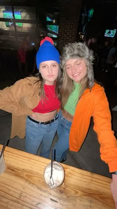 two women standing next to each other at a table with a drink in front of them