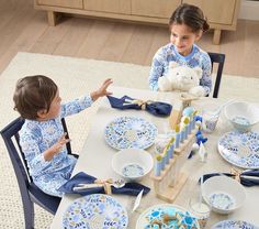 two children sitting at a table with blue and white dishes on it, one holding a teddy bear