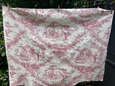 a pink and white toiler print fabric hanging on a clothes line in front of some bushes