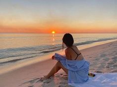 a woman sitting on top of a sandy beach next to the ocean at sunset or dawn