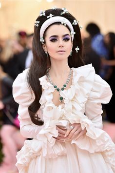 a woman with long dark hair wearing a white dress and headpiece, standing in front of a crowd