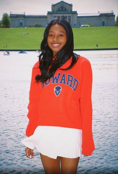 a woman standing in front of a body of water wearing an orange shirt and white skirt