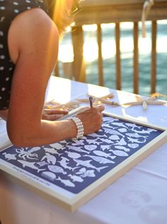 a woman writing on a piece of paper while sitting at a table with water in the background