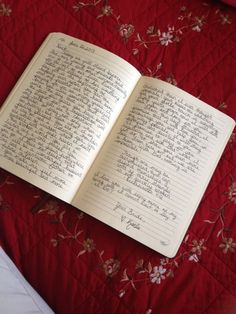 an open notebook sitting on top of a red bed spreaded with flowers and writing