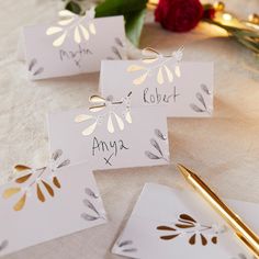 three place cards with gold foil on them sitting on a table next to flowers and a pen