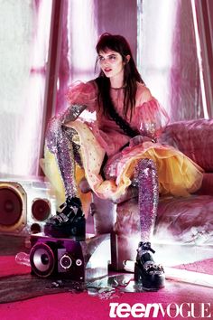 a woman sitting on top of a pink couch next to a pair of boots and a camera