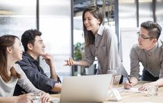 four people sitting at a table in front of a laptop computer and talking to each other