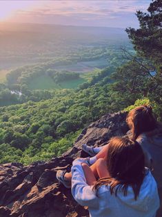 two people sitting on the edge of a cliff looking out over a valley and forest