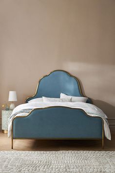 a blue bed with white sheets and pillows in front of a beige wall, next to a lamp