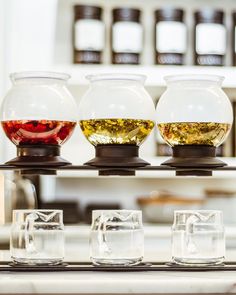 four glasses filled with different types of tea on a tray in front of shelves full of jars