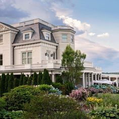 a large white house surrounded by trees and flowers on a cloudy day with blue skies in the background