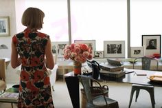 a woman standing in an office looking out the window at her work desk and flowers