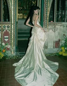 a woman in a long white dress is standing by a wall with arches and flowers