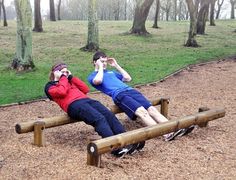 two people sitting on a wooden bench in the park, one is looking through binoculars