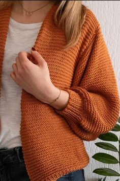 a woman wearing an orange knitted cardigan and jeans with her hands folded in front of her chest