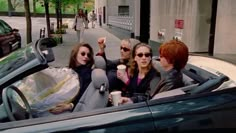 three women sitting in the back seat of a convertible car and one woman holding a coffee cup