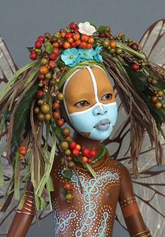 a woman with painted face and body is standing in front of a gray background, wearing an elaborate headdress