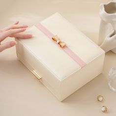 a white box with a pink ribbon on the top and two gold rings next to it