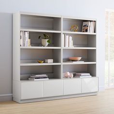 a white bookcase with books and magazines on it next to a window in an empty room