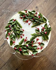 a cake decorated with green leaves and red berries on a clear plate sitting on top of a wooden table