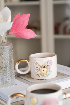 a pink flower sitting on top of a table next to two coffee mugs and a book